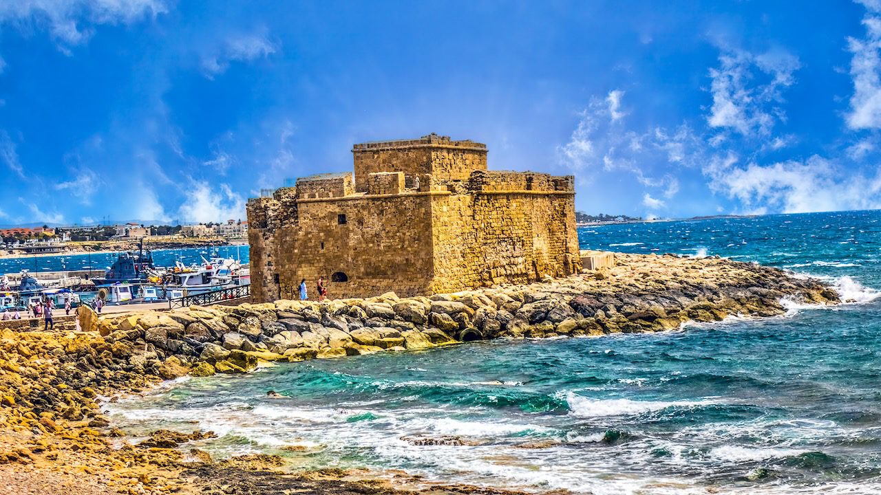 Pafos Castle, a historical fortress by the sea in Cyprus, under a bright blue sky with waves crashing against the rocky shore.