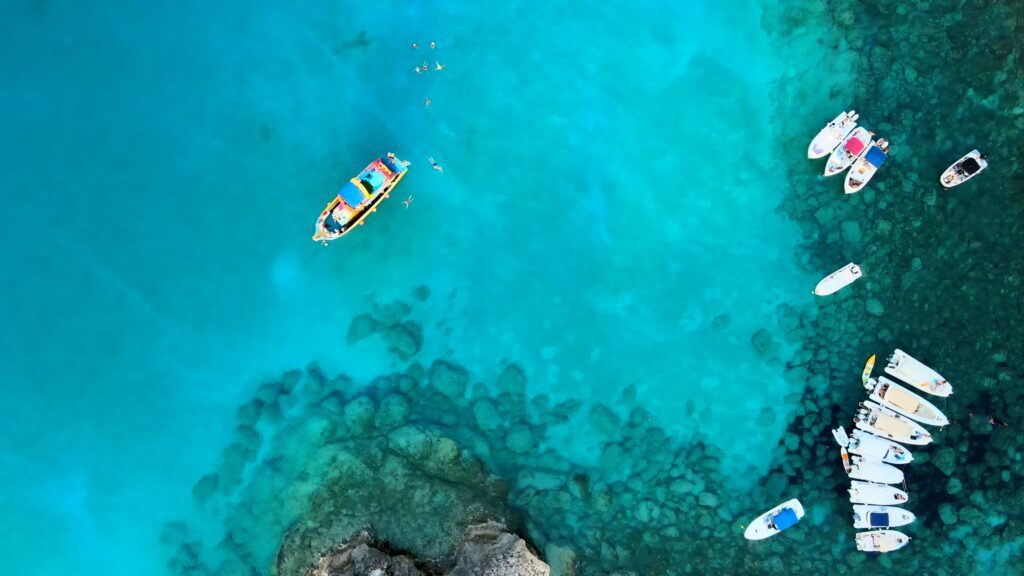 Drone view of boats anchored in the natural bay at Cavo Greco, with swimmers enjoying the crystal clear blue waters, highlighting the serene and vibrant aquatic landscape.