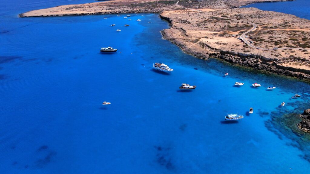 Aerial shot of Cape Greco's bustling Blue Lagoon in summer: boats scattered amidst shimmering azure waters.