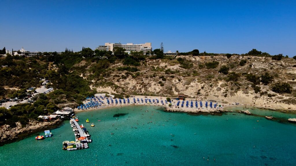 Aerial view of Konnos Bay, showcasing its clear waters, sandy beach, and the Grecian Park Hotel atop the cliff.