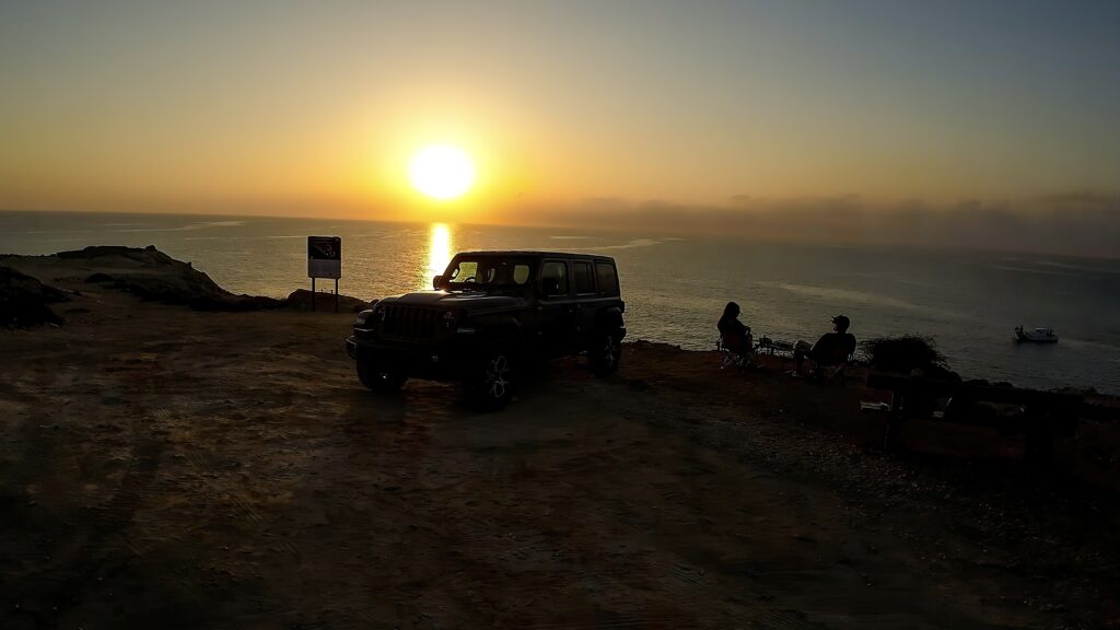 Two people sitting at the edge of a cliff at Cavo Greco, watching the sunrise with their jeep parked nearby. The man wears goggles, a detail that hints at his earlier flight with an FPV drone.