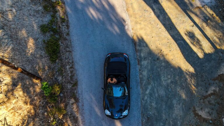 Aerial view of a man driving a convertible sports car on a narrow, winding road through the Troodos Mountains, Cyprus, with shadows of trees stretching across the road.