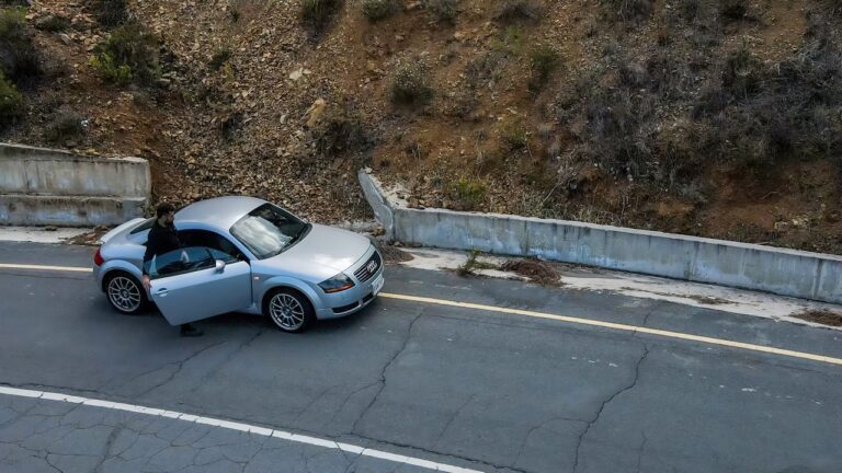 A man enters his silver Audi TT on a winding mountain road in the Troodos Mountains, Cyprus, on a chilly day, with a background of rocky terrain and sparse vegetation.